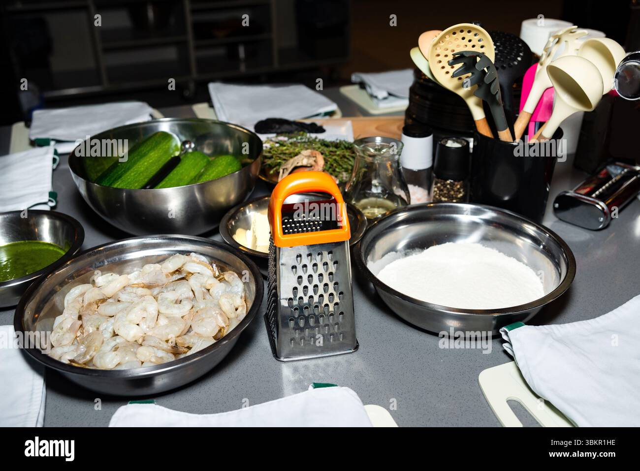 Un ripiano della cucina con gamberi freschi, zucchine, farina, erbe e utensili da cucina preparati per una sessione di cucina gourmet. Foto Stock