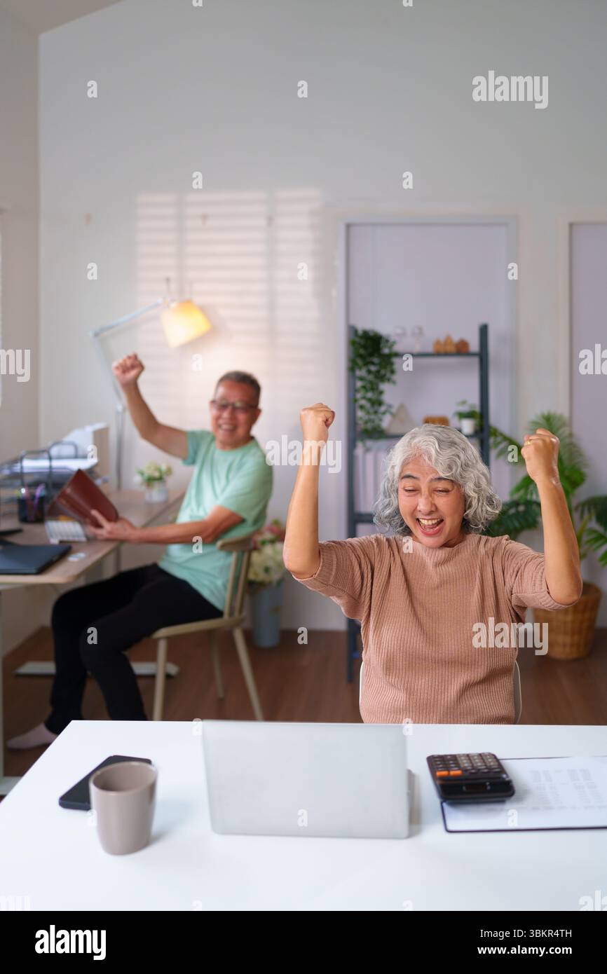 Una felice coppia asiatica più anziana che festeggia un successo a casa con laptop e documenti. Raggiungono gli obiettivi insieme gestendo i loro fondi pensionistici. Foto Stock
