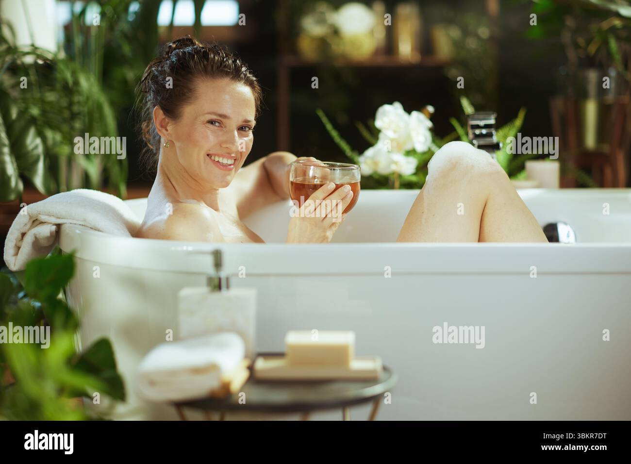 Una donna sorridente di mezza età gode di un lussuoso bagno di bolle, che tiene il tè. Capelli bagnati tirati indietro, si rilassa tra lussureggianti piante verdi e luci calde, e. Foto Stock