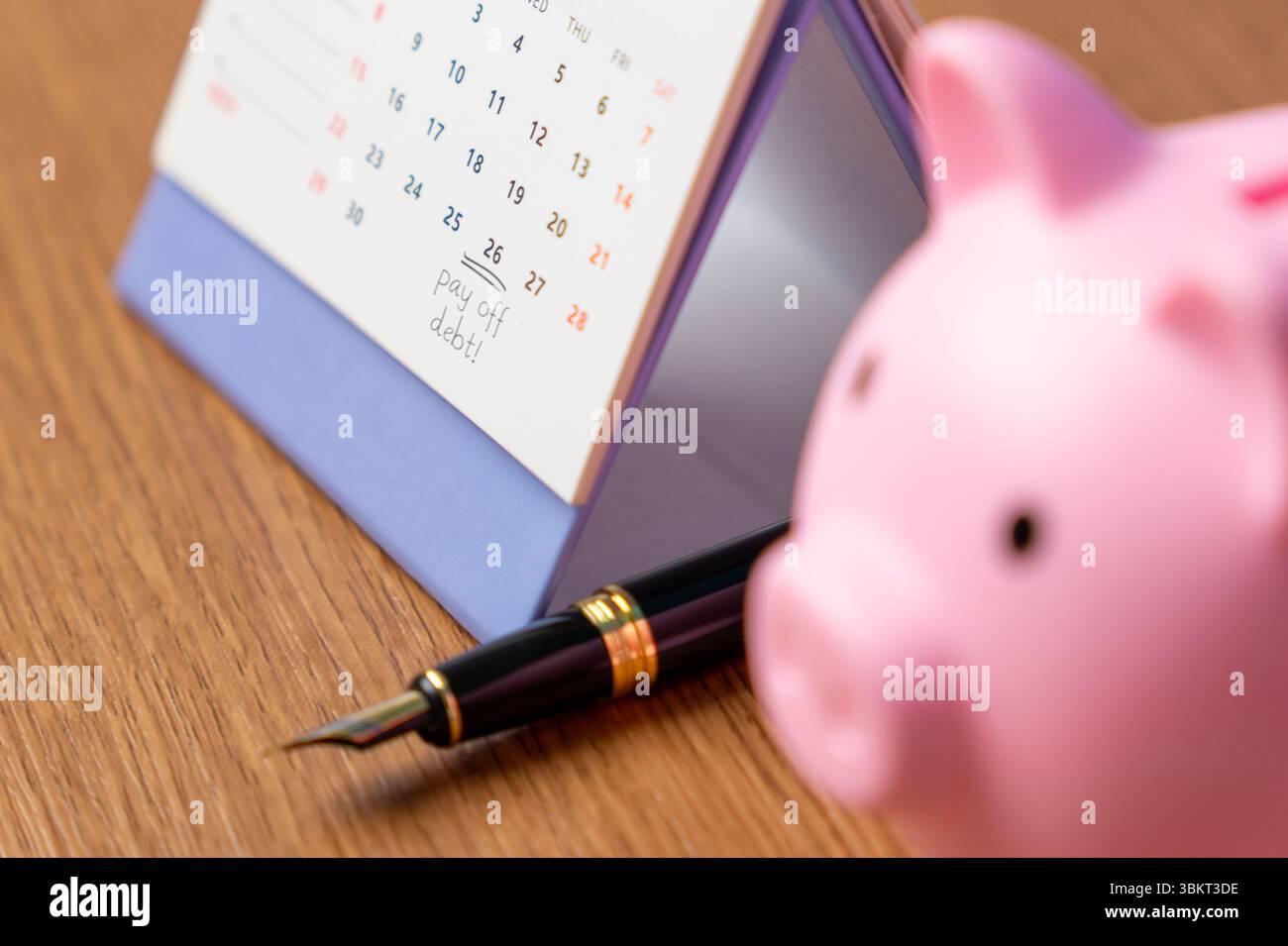 Un calendario da scrivania con "pagare il debito!" segnata su una data accanto a una penna e a un salvadanaio. Pianificazione finanziaria, concetto di gestione del debito. Foto Stock
