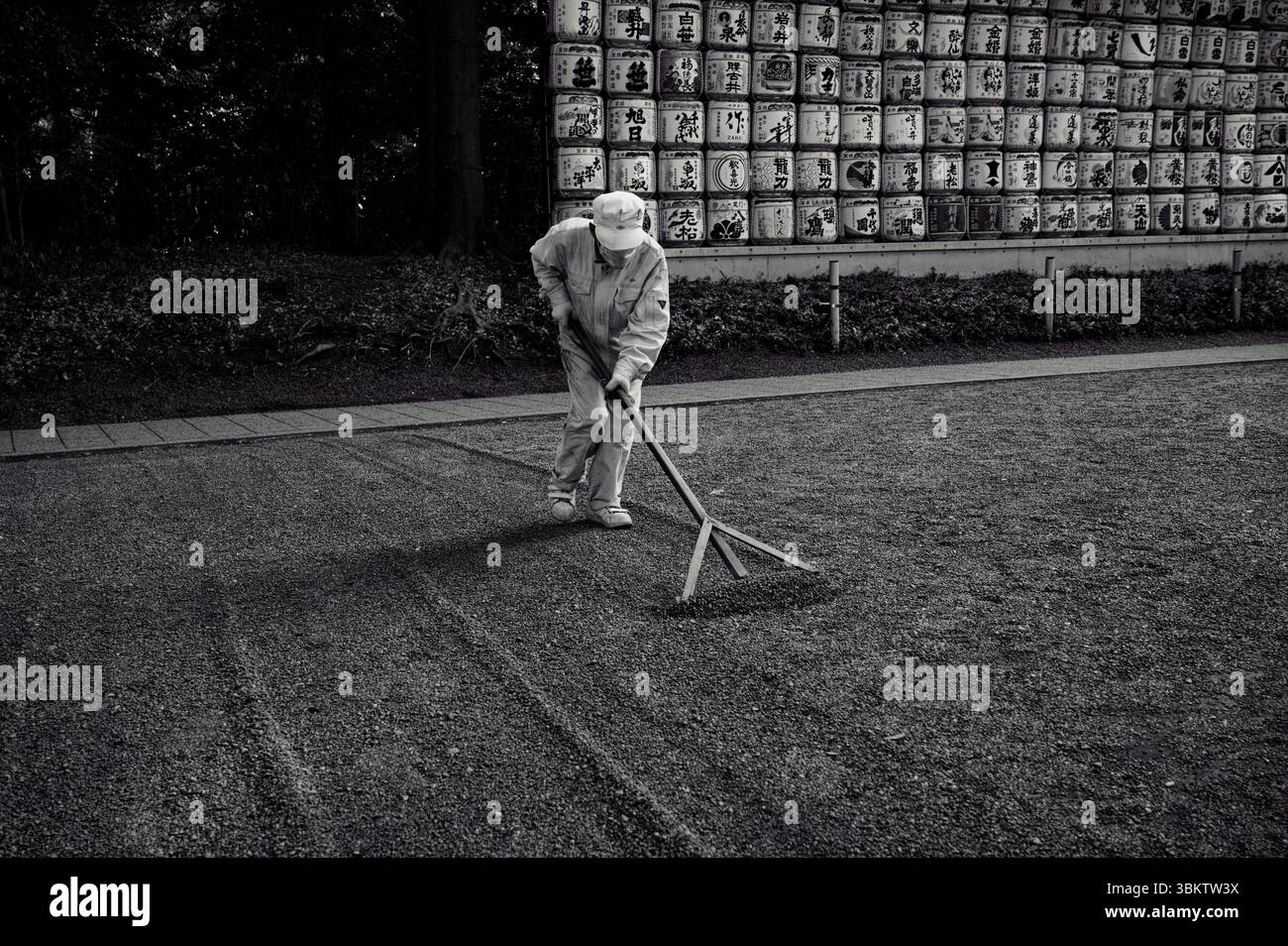 Il lavoratore del Santuario rastrella con cura la ghiaia davanti a una parete di botti decorative di sake, mantenendo il terreno con la massima precisione Foto Stock