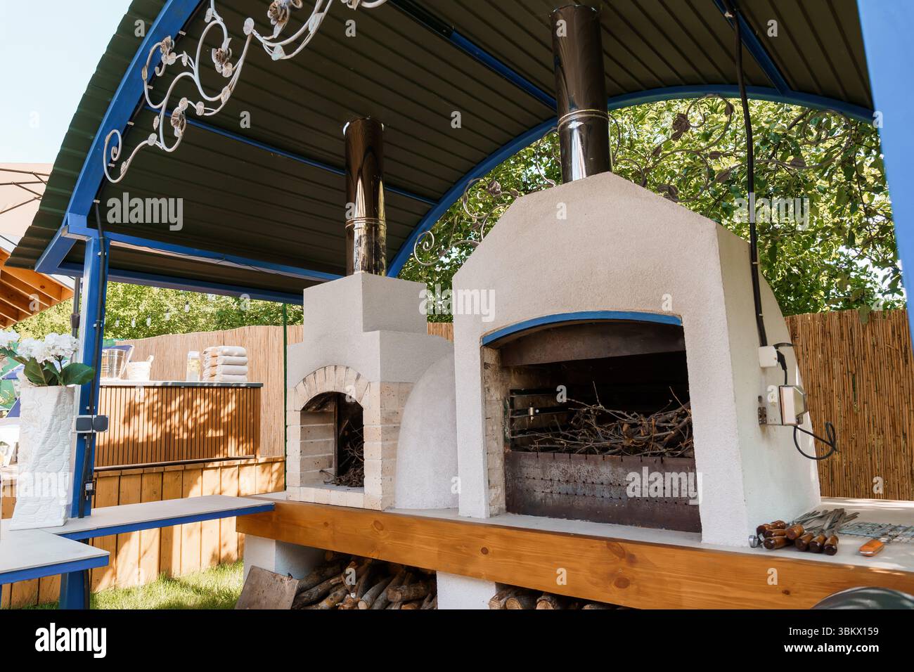 Un'accogliente cucina all'aperto presenta un forno a legna in pietra integrato con un fumatore, coperto sotto un pergolato blu, che mostra legno e utensili da cucina Foto Stock Un'accogliente cucina all'aperto presenta un forno a legna in pietra integrato con un fumatore, coperto sotto un pergolato blu, che mostra legno e utensili da cucina Foto Stock