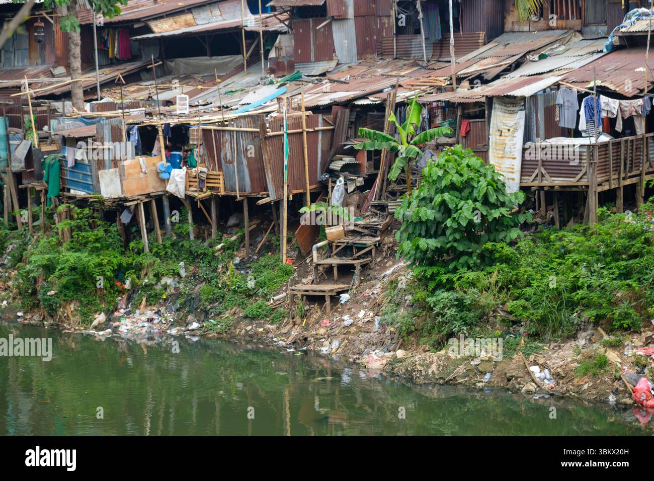 Le case improvvisate fiancheggiano una sponda inquinata del fiume, dove vivono i residenti e asciugano i vestiti in mezzo alla spazzatura. Foto Stock