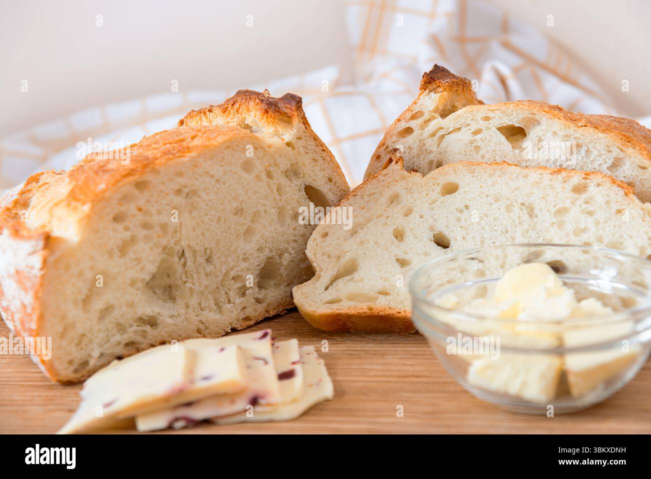 La pagnotta affettata di pane croccante si trova su una superficie di legno accanto a una ciotola di burro cremoso e fette di formaggio, creando un'atmosfera calda. Foto Stock
