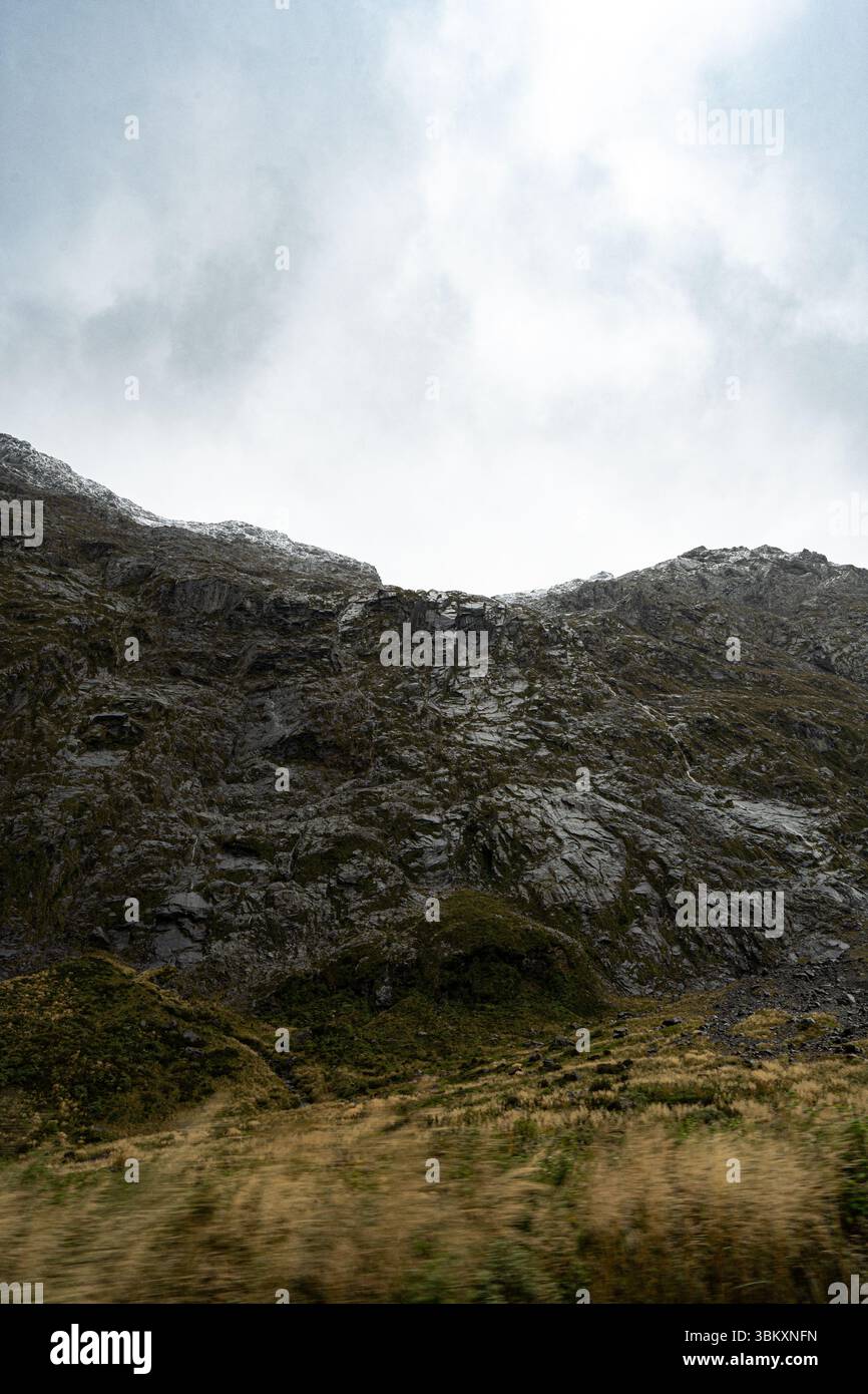 Un'aspra montagna si erge ripidamente sotto un cielo torbido e coperto. Rocce scure e frastagliate e macchie di vegetazione verde dominano il primo piano, evocando un Foto Stock