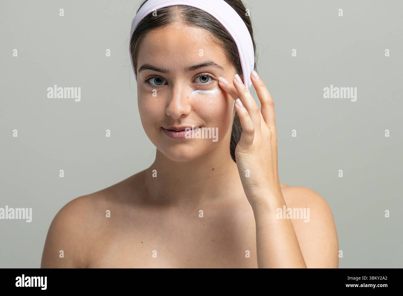 Una bella giovane donna guarda la fotocamera mentre applica delicatamente una crema nutriente per gli occhi. Concetto di cura della pelle anti-invecchiamento e trattamento dei cerchi scuri Foto Stock