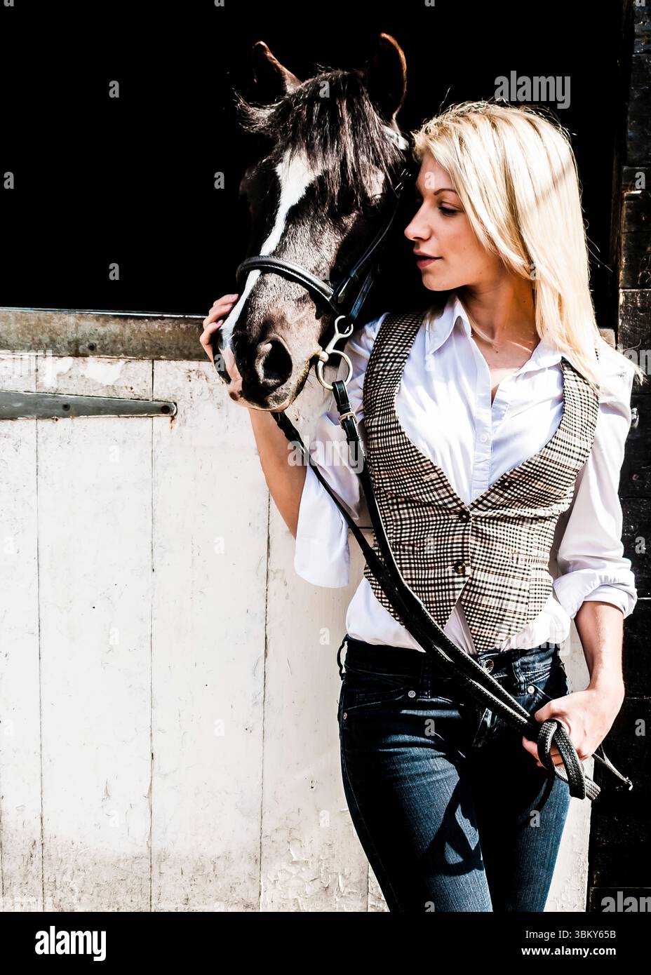 Una donna con una camicia bianca e un giubbotto è in piedi accanto a un cavallo, tenendo le redini. Foto Stock