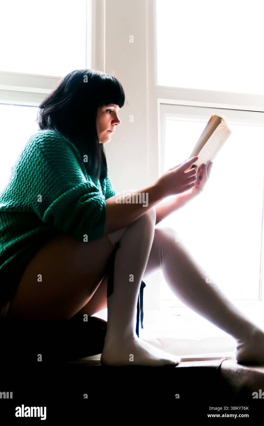 Una donna con i capelli neri è seduta su un davanzale, a leggere un libro. Indossa un maglione verde e calze bianche. Foto Stock