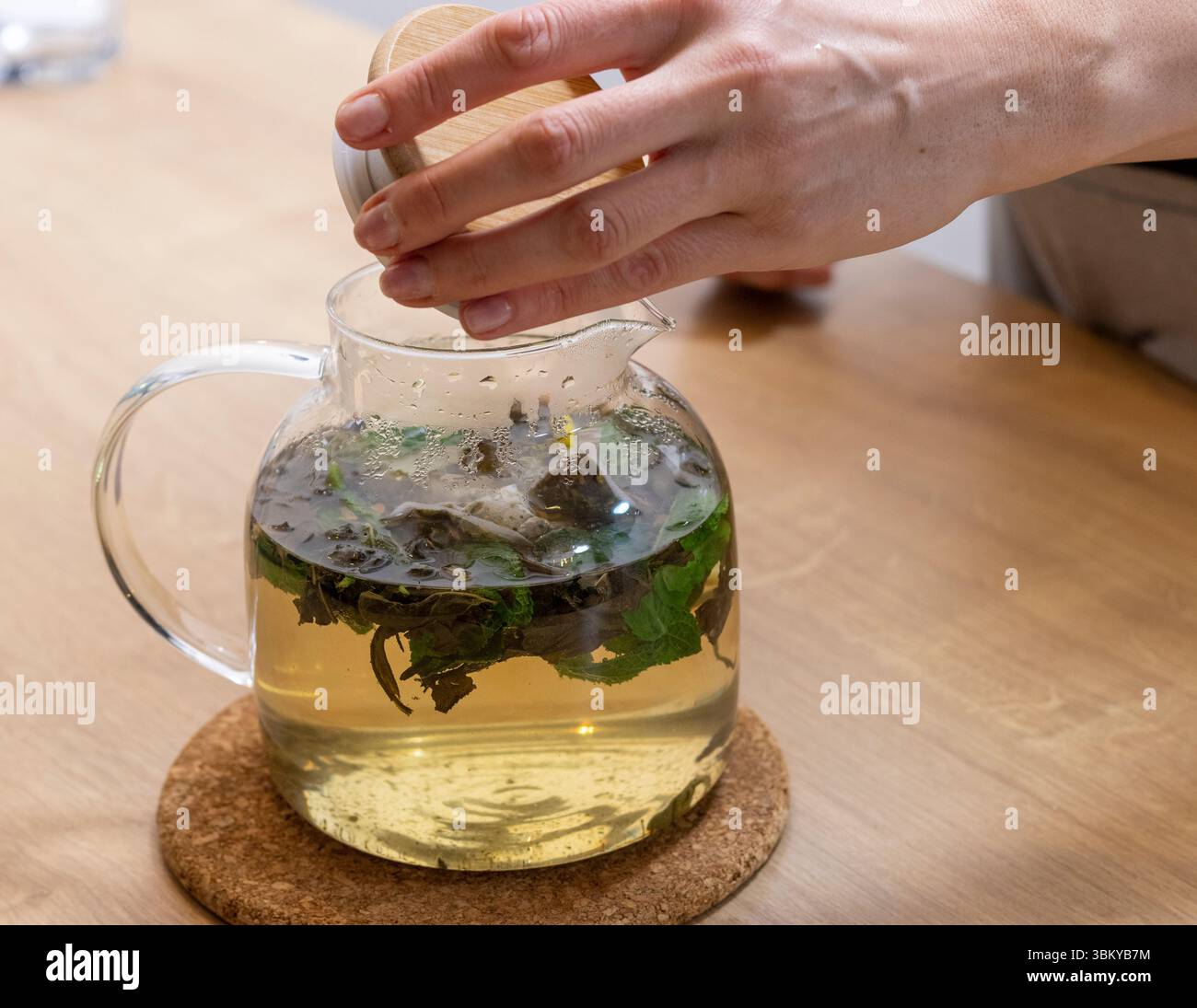 Donna apre il coperchio in legno dell'elegante teiera trasparente con foglie di tè verde appena preparate, foglie di menta e fette di limone Foto Stock