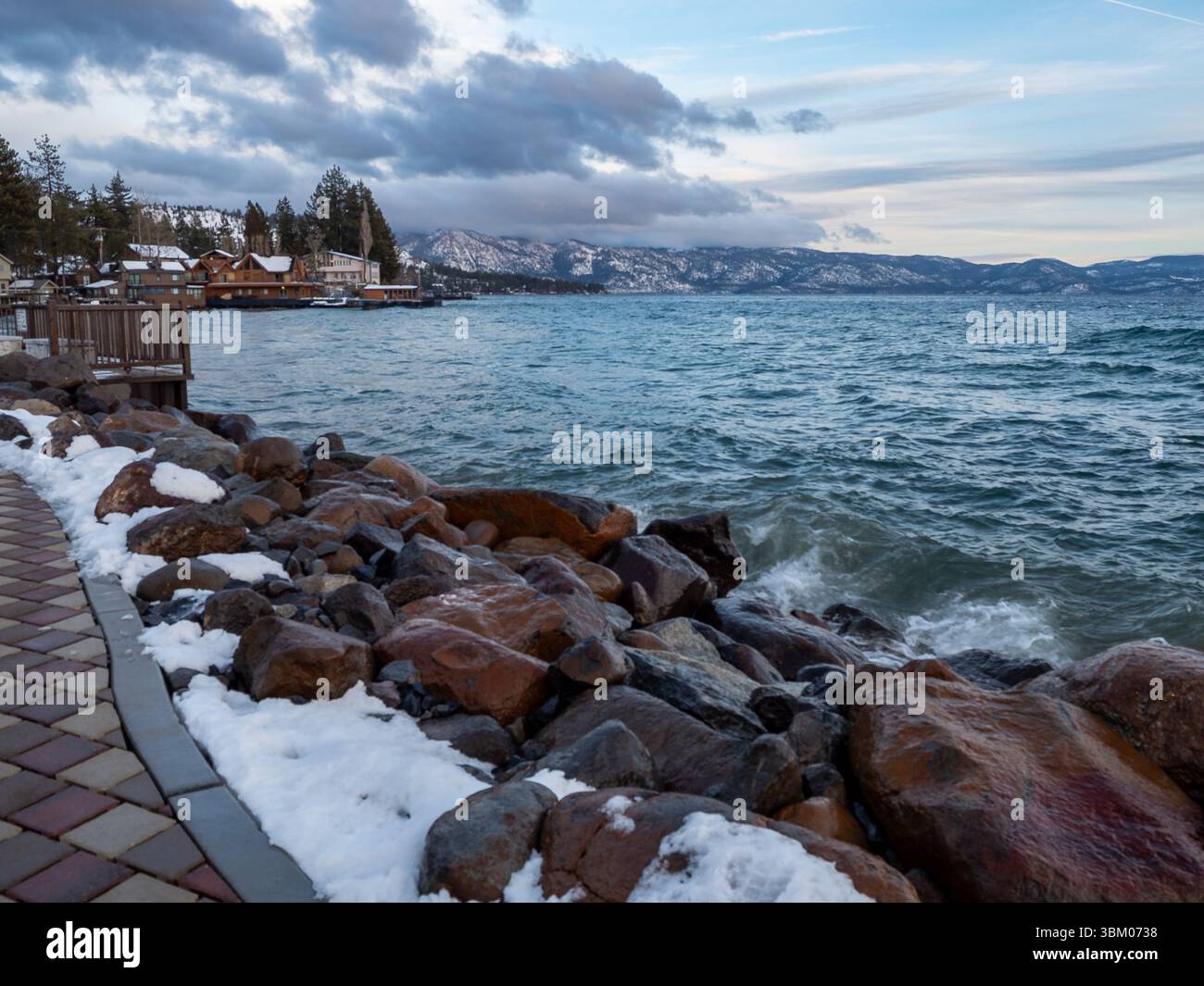 Una vista panoramica di una costa rocciosa con macchie di neve, dove le onde si infrangono contro le rocce. Sullo sfondo, ci sono case e alberi lungo la costa, con montagne visibili in lontananza sotto un cielo nuvoloso. Foto Stock