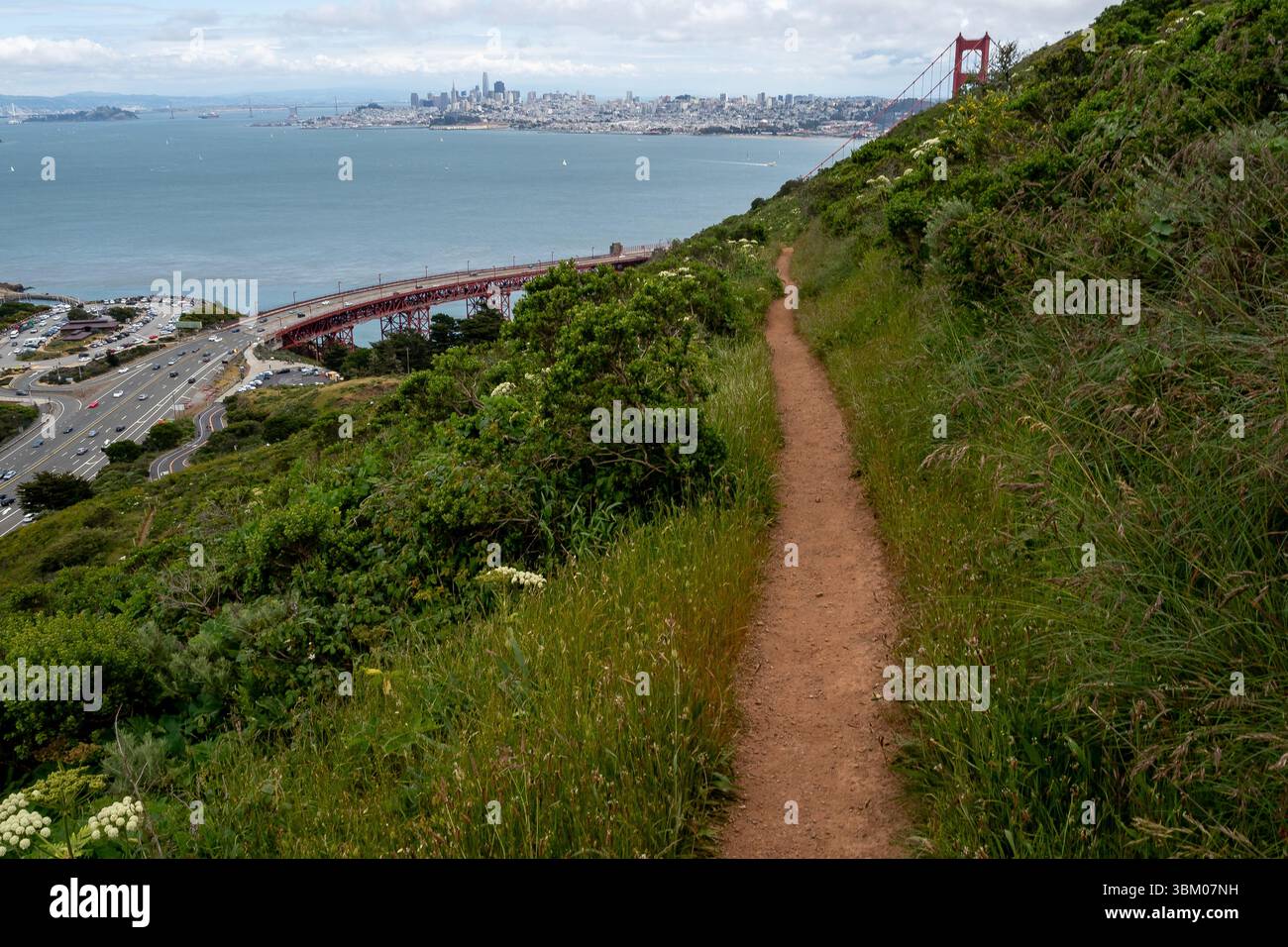 Un sentiero sterrato si snoda lungo una collina, offrendo una vista panoramica del Golden Gate Bridge e dello skyline di San Francisco in lontananza, invitando a esplorare e apprezzare la bellezza naturale e i monumenti iconici. Foto Stock
