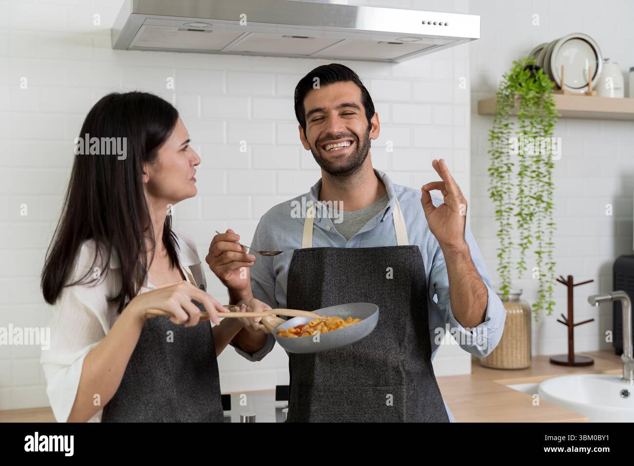 Una giovane coppia prepara il pasto in cucina. L'uomo assaggia il cibo dal cucchiaio e mostra un buon segno con un sorriso gioioso. La donna tiene in mano la padella. Stile di vita domestico moderno Foto Stock