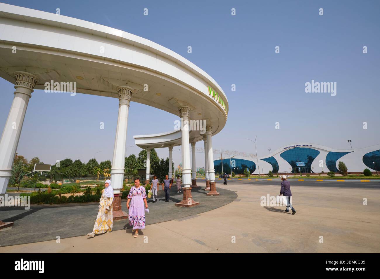 Vista del vicino parco con l'edificio moderno dietro di esso. Al moderno e curvato centro sportivo Megasportkomplex di Qarshi, Uzbekistan. Foto Stock