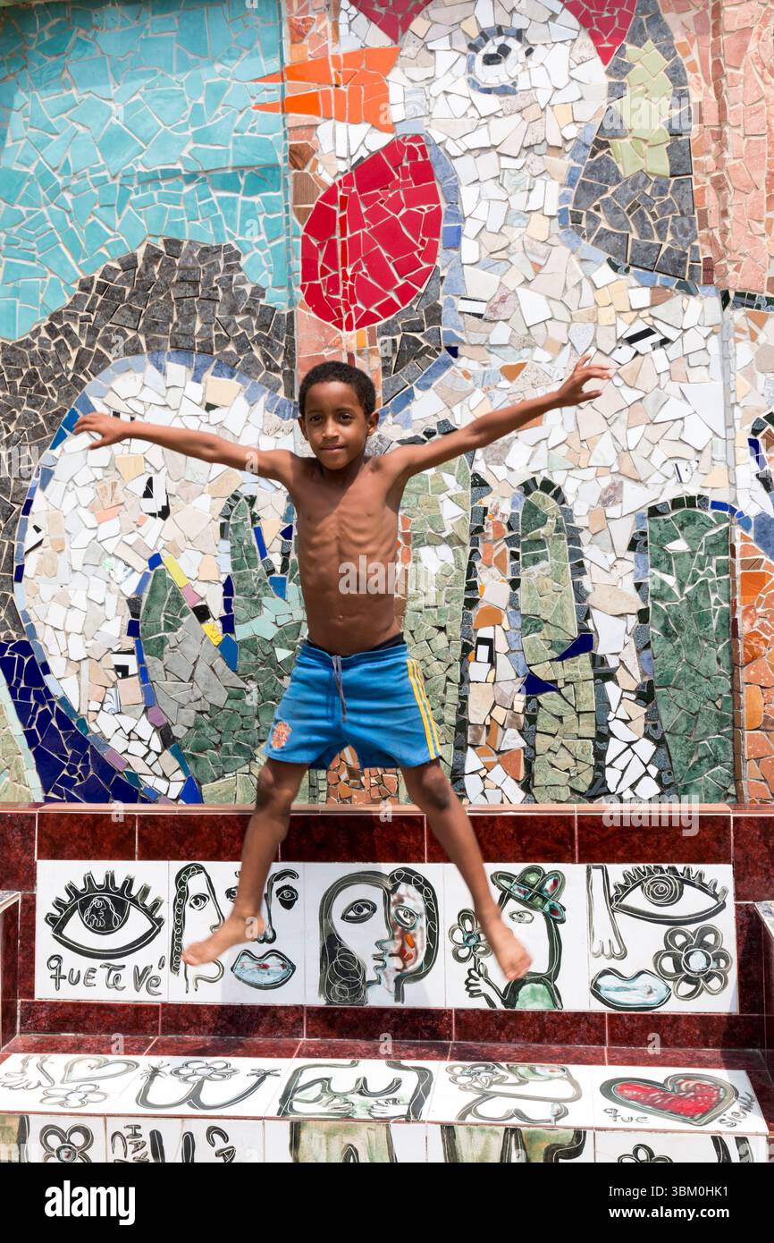 Cuba, l'Avana. Jaimanitas. Arte, mosaico, casa di Jose Fuster. Murales e mosaici decorati. Ragazzo che salta di fronte al murale a mosaico. (Solo per uso editoriale) Foto Stock