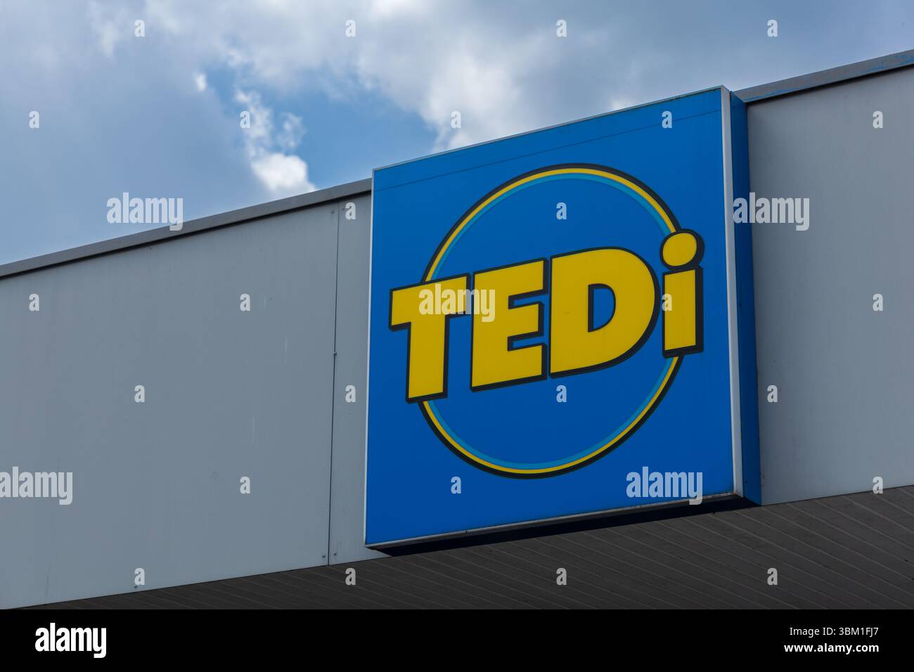 Un cartello blu Tedi per la vendita al dettaglio con scritta gialla e design circolare montato sull'esterno di un edificio grigio. Kleve, Germania. 22 aprile 2025. Foto Stock