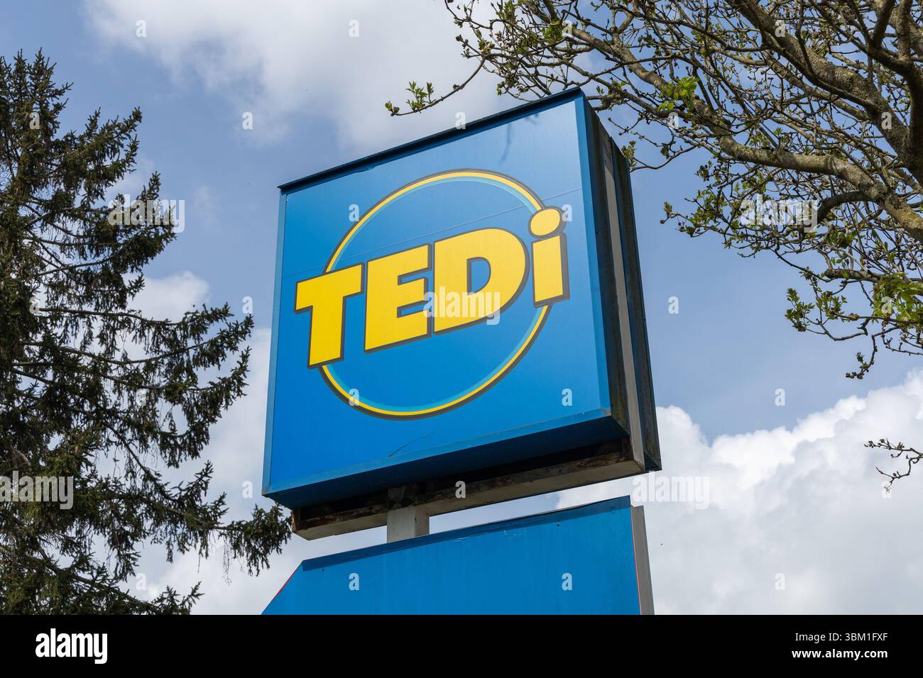 Un cartello blu luminoso del negozio Tedi con scritta gialla e un design circolare si staglia contro un cielo parzialmente nuvoloso. Kleve, Germania. 22 aprile 2025. Foto Stock