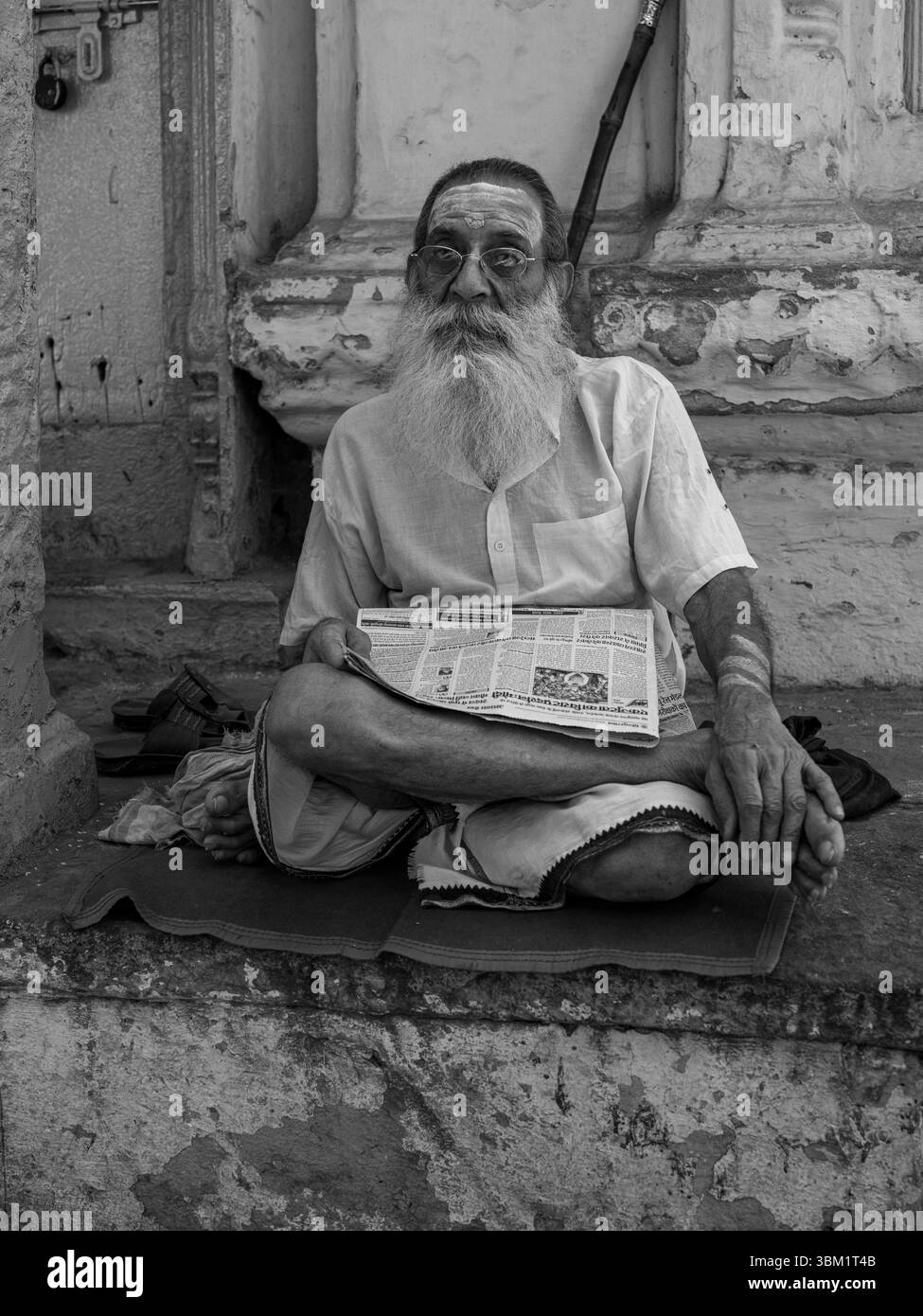 India, Varanasi, 16 marzo 2025. Uomo anziano con barba lunga e occhiali seduto a gambe incrociate sui gradini del tempio a leggere un giornale. Ritratto monocromatico di da Foto Stock