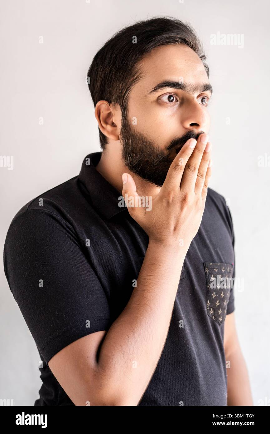 Una fotografia ravvicinata mostra un uomo con la barba che indossa una polo nera. La sua espressione e' di sorpresa o shock, la sua mano che copre la bocca. T Foto Stock