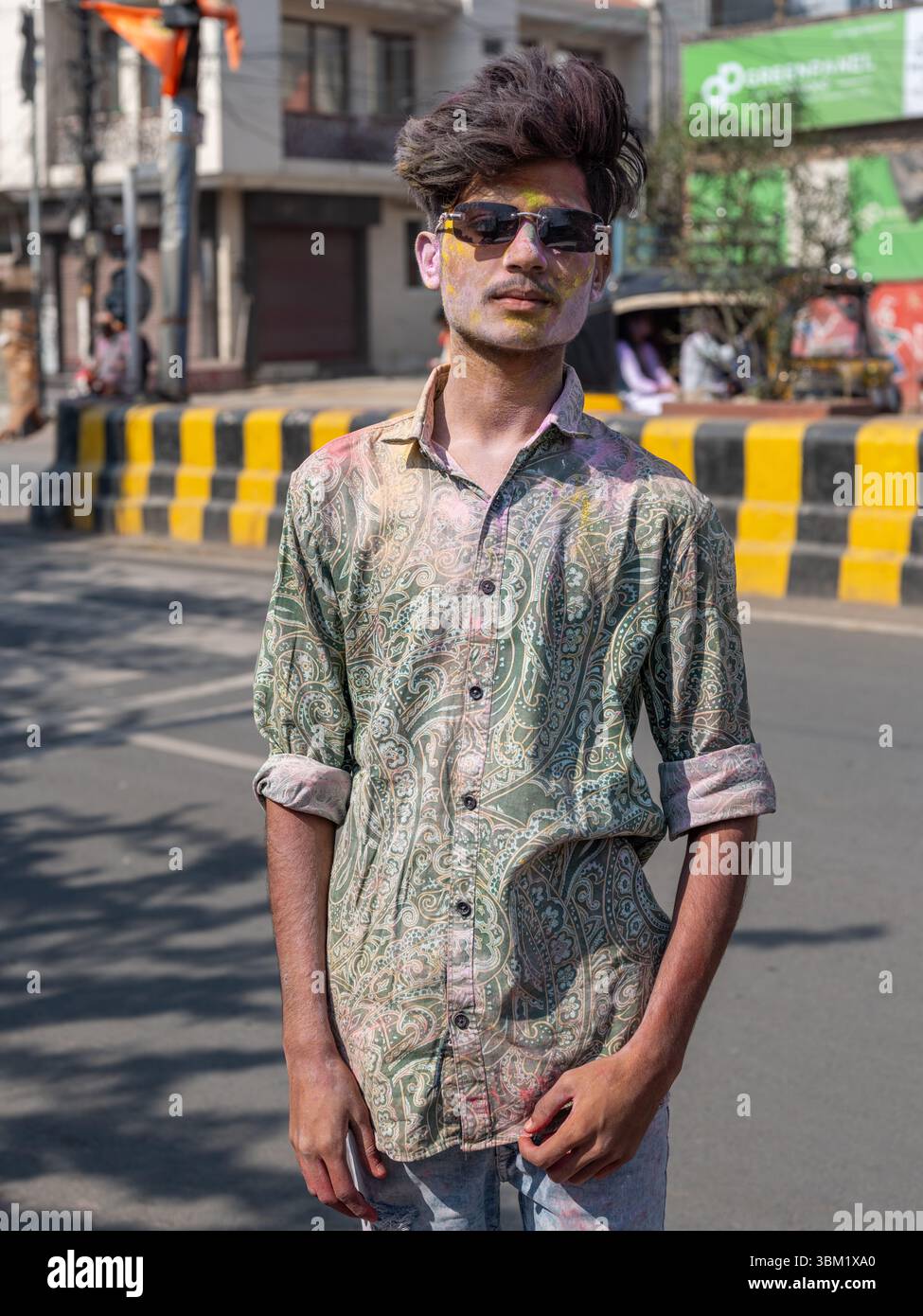 India, Rajasthan, 22 marzo 2025. Giovane indiano con capelli e occhiali da sole eleganti, viso ricoperto di colori Holi, che posa durante il festival Holi in città Foto Stock