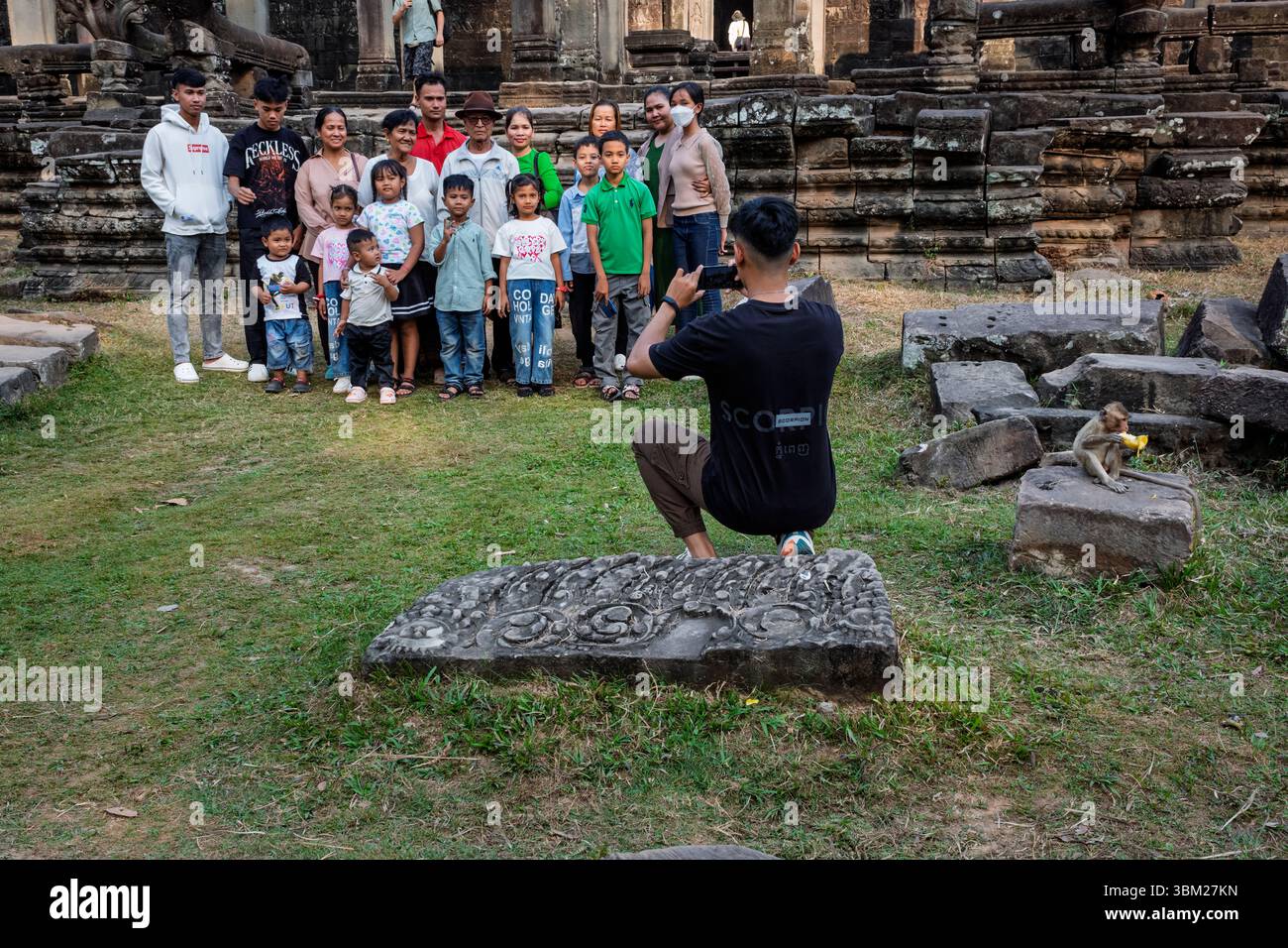 Una posa in famiglia per una foto di gruppo mentre un macaco mangia una banana accanto a loro, Bayon Temple, Angkor Thom, Siem Reap, Cambogia Foto Stock