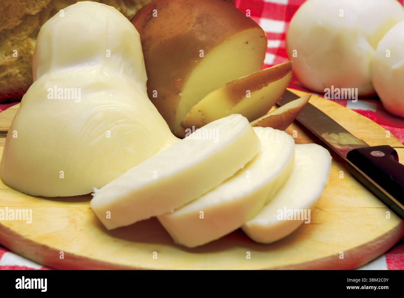 Scamorza a fette con provola intera e caciocavallo poggiati su un tagliere di legno con pane e coltello su una linguetta a scacchi rossa e bianca Foto Stock