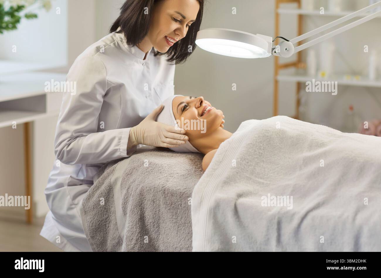 Cosmetologo che esamina il viso di giovane donna sorridente sotto lampada prima di iniziare la procedura di cura della pelle. Foto Stock