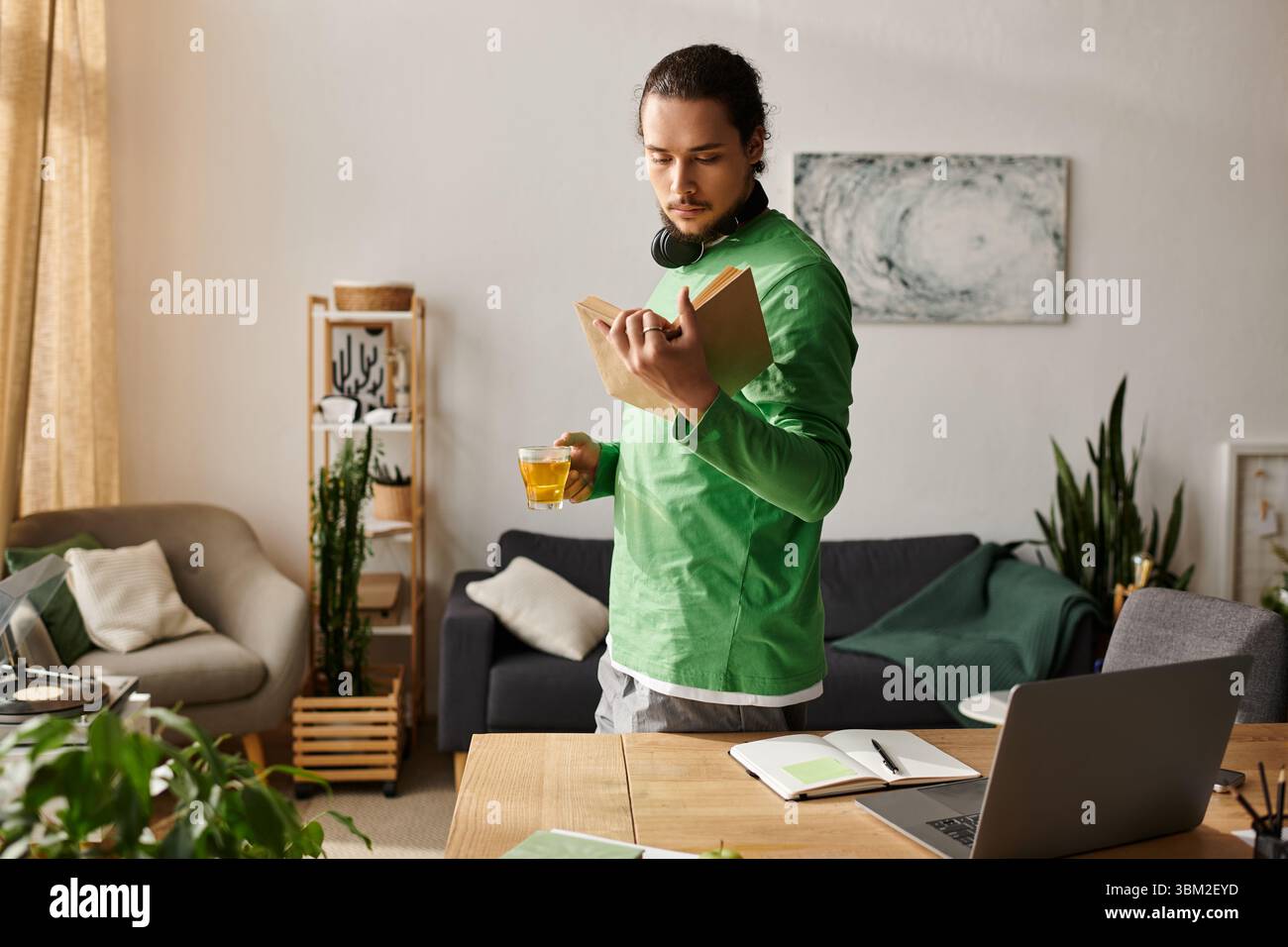 Un giovane si trova in un accogliente soggiorno, assorbito in un libro con un drink in mano. Foto Stock