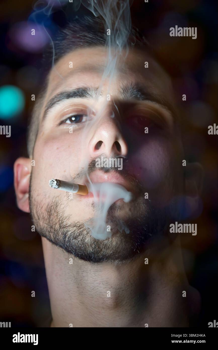 Un ritratto ravvicinato di un uomo che espira fumo da una sigaretta, catturando un'espressione intensa su uno sfondo colorato e sfocato. Foto Stock