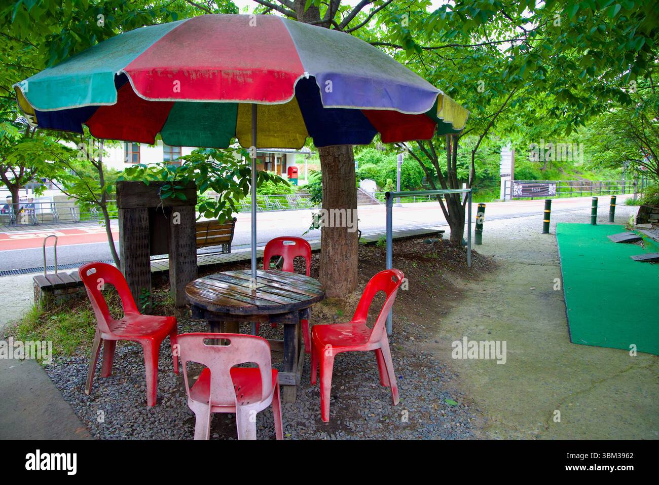 Namyangju, Corea del Sud - 19 maggio 2025: Un tavolo da patio intempestivo con sedie di plastica rossa e un ombrello multicolore offre una sosta informale per riposare in bicicletta Foto Stock