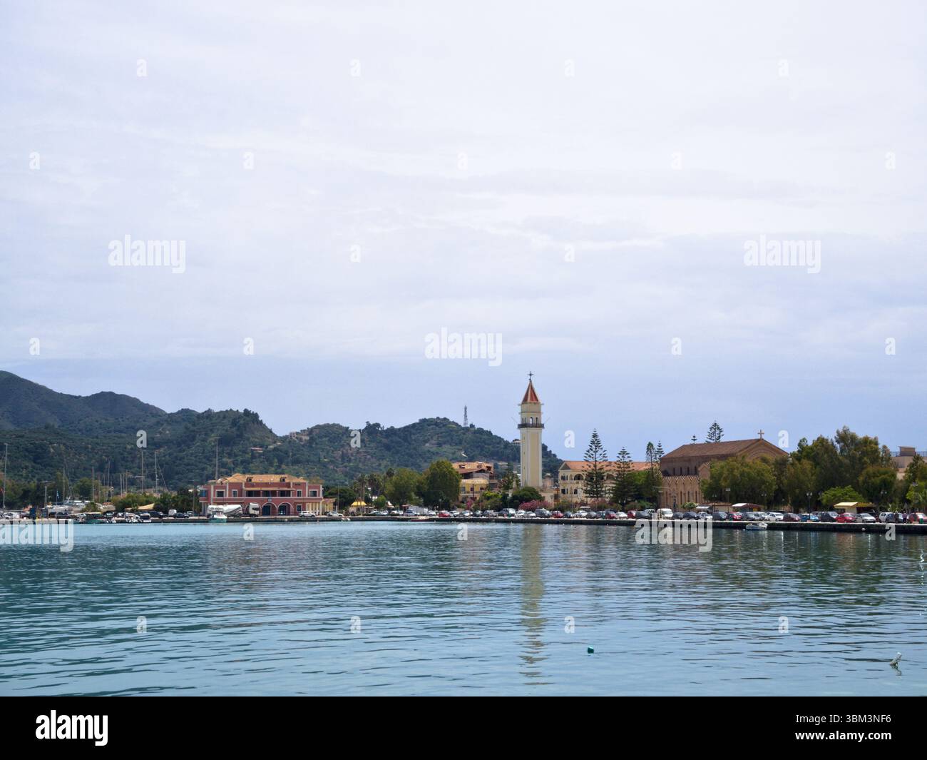 La città di Zante è vista dal porto con il mare calmo, la torre della chiesa centrale, lo sfondo della collina e il cielo mediterraneo nuvoloso. Foto Stock