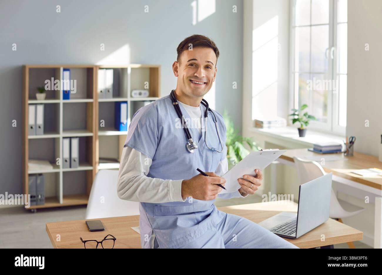 Ritratto di un giovane medico maschio felice in uniforme medica blu seduta alla scrivania nel suo ufficio. Foto Stock