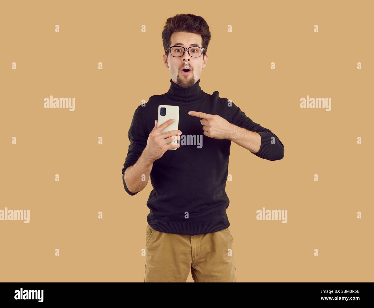 Il giovane brunet con dolcevita nero è stupito e punta lo smartphone su sfondo beige. Foto Stock