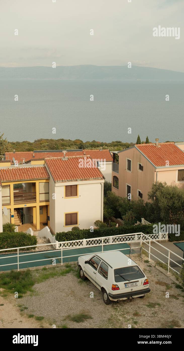 Un'antica Volkswagen bianca poggia su un'area di ghiaia accanto a un gruppo di case color pastello mediterranee. I loro tetti in terracotta. Foto Stock