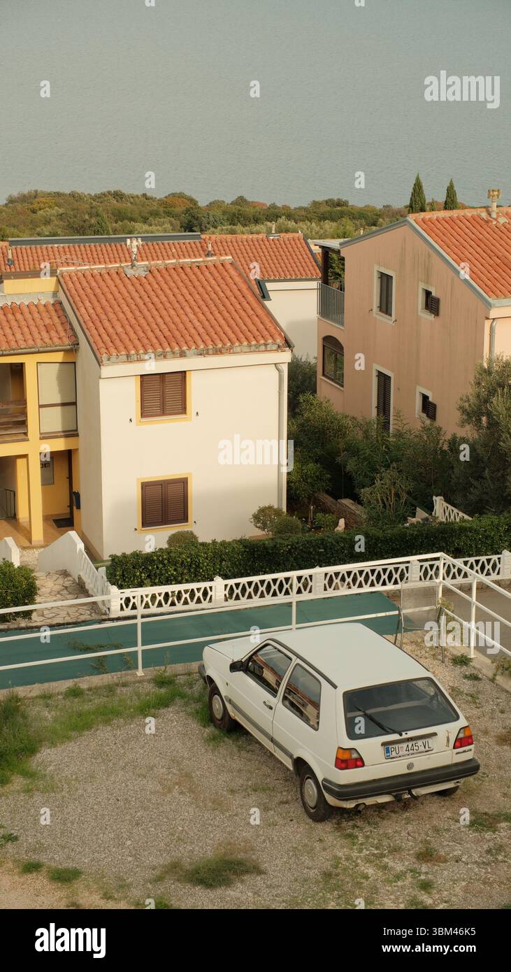 Un'antica Volkswagen bianca poggia su un'area di ghiaia accanto a un gruppo di case color pastello mediterranee. I loro tetti in terracotta. Foto Stock