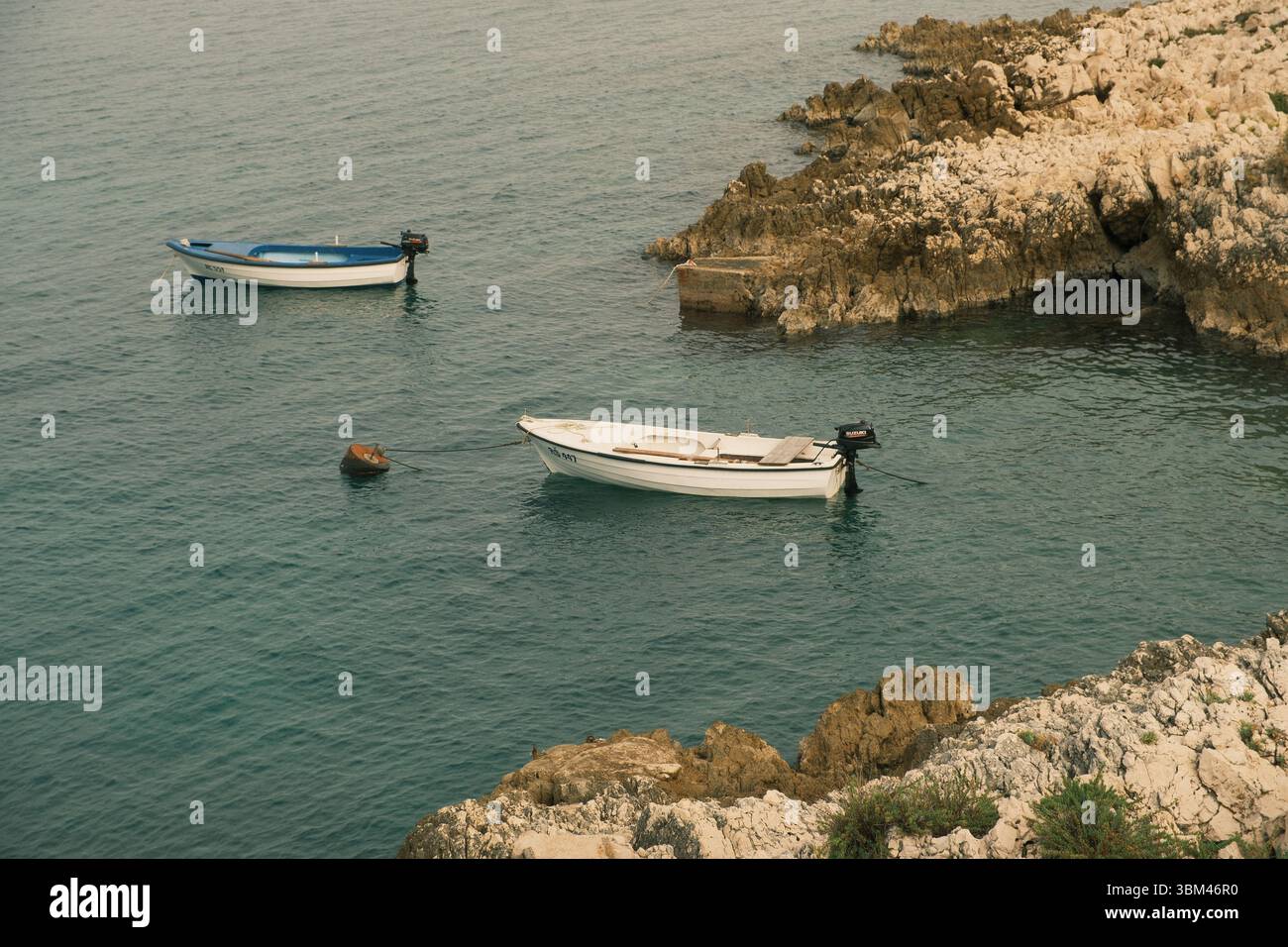 Due piccole barche riposano tranquillamente in un'isolata insenatura adriatica, ancorata vicino a un'aspra costa rocciosa. L'acqua è limpida e ferma, tenendo delicatamente i recipienti. Foto Stock