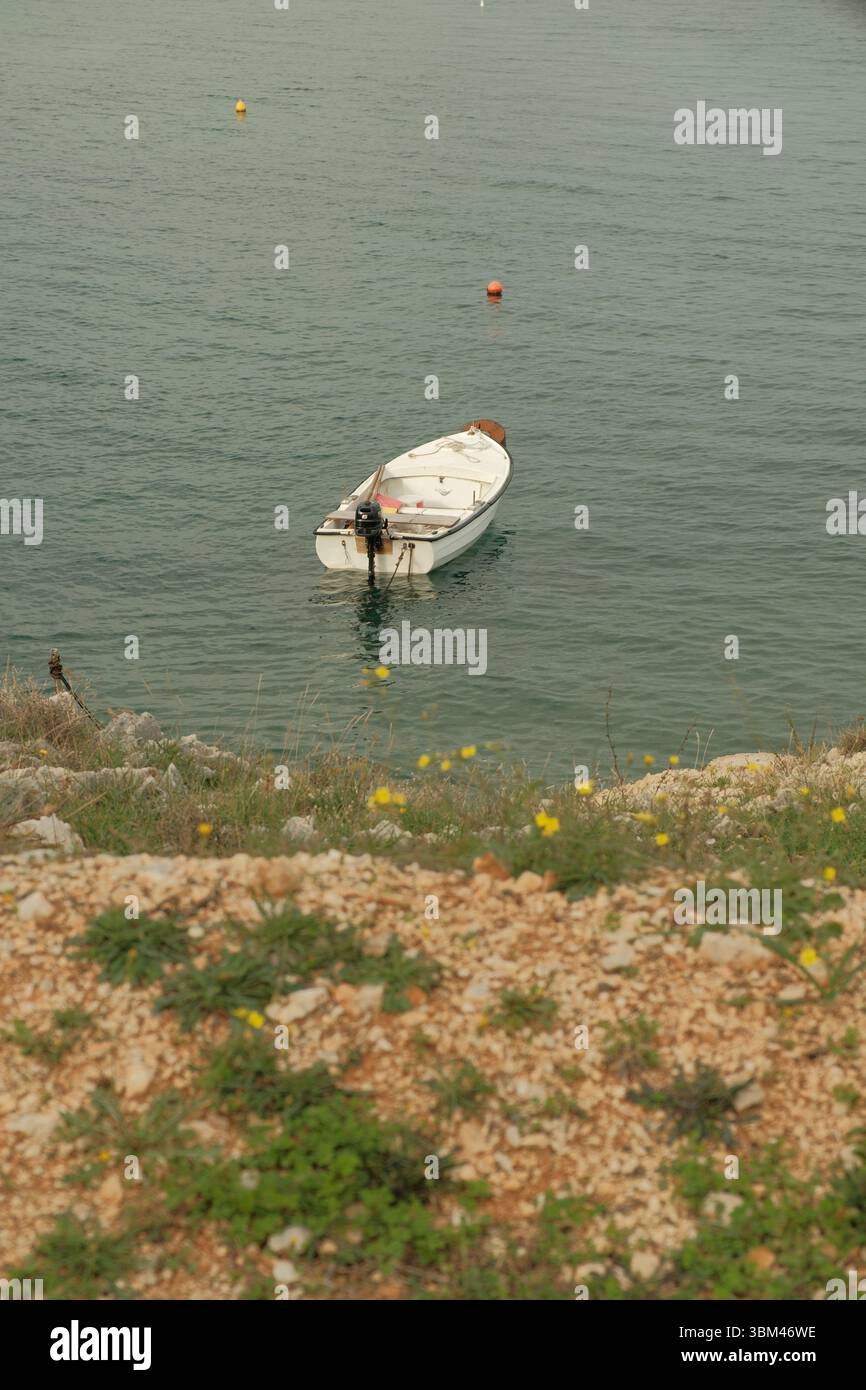 Una barca bianca solitaria galleggia dolcemente sul tranquillo mare blu-verdastro, ancorata vicino alla costa rocciosa e erbosa. Foto Stock