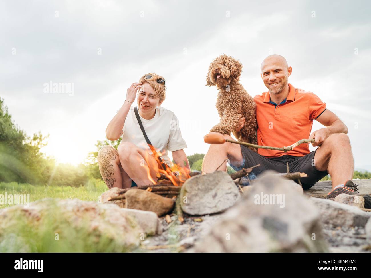 Graziosa scena di picnic all'aperto: Coppia felice sorridente con il cane Maltipoo che grigia salsicce su bastoncini da barbecue seduti accanto al fuoco durante il tour di trekking Foto Stock