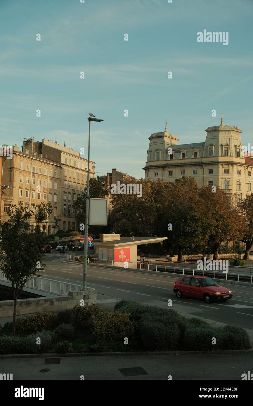 Una leggera luce mattutina tocca le facciate color pastello di una vecchia strada europea, dove una solitaria auto rossa attraversa la tranquilla scena urbana. Foto Stock