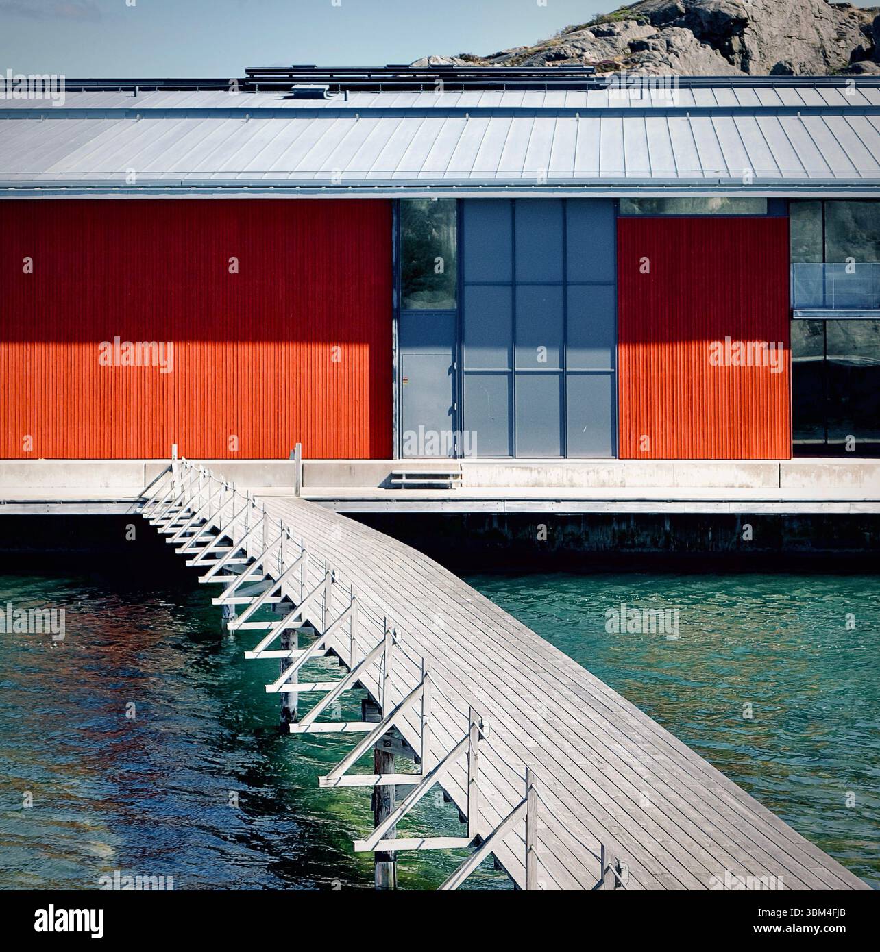 Edificio dal design dipinto di rosso a contrasto con elementi minimalisti contro l'isola rocciosa con ponte sulle acque del mare Foto Stock