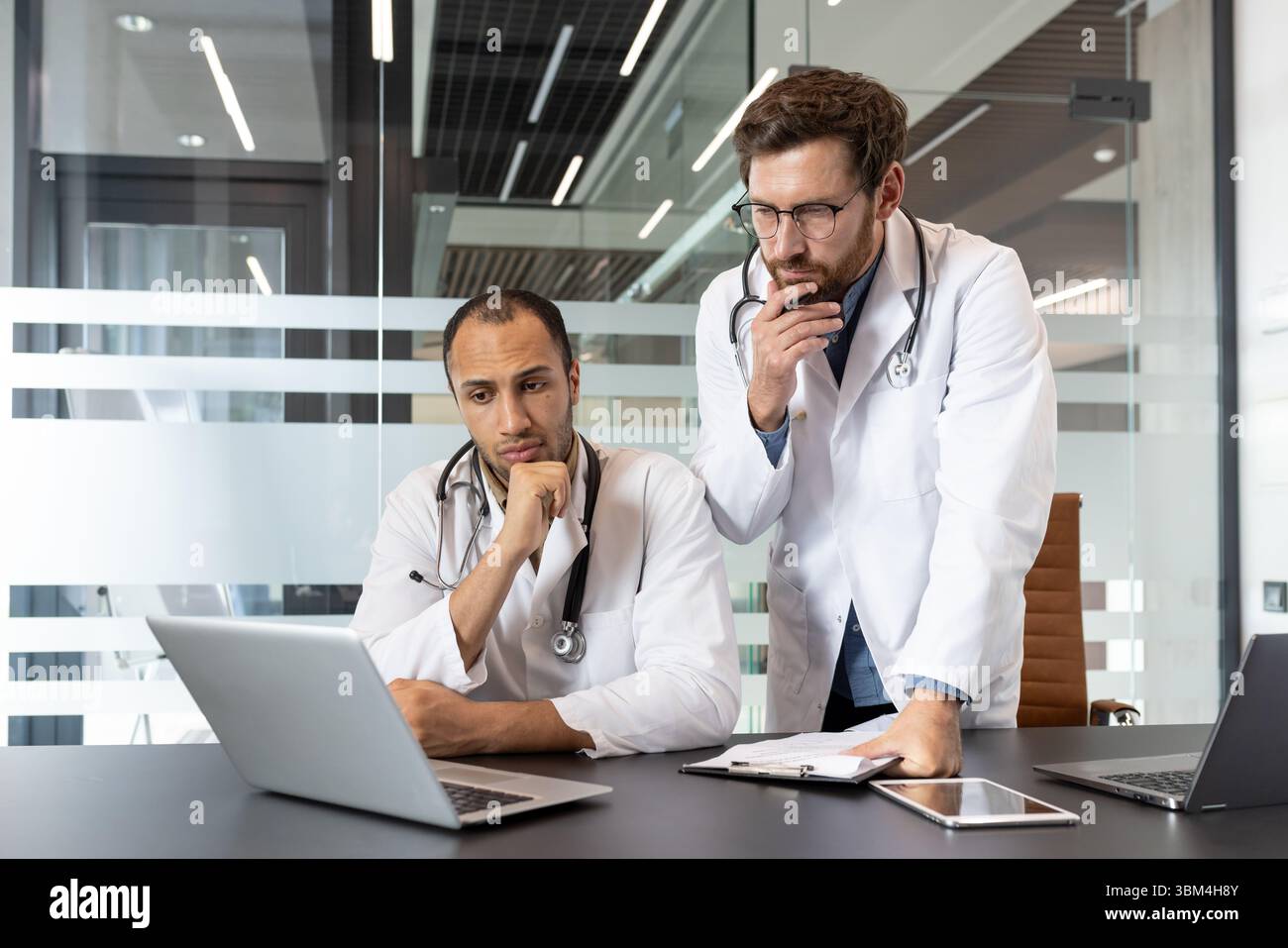 Due medici, che indossano cappotti e stetoscopi bianchi, guardano lo schermo di un notebook in un ufficio moderno, con una profonda riflessione. Foto Stock