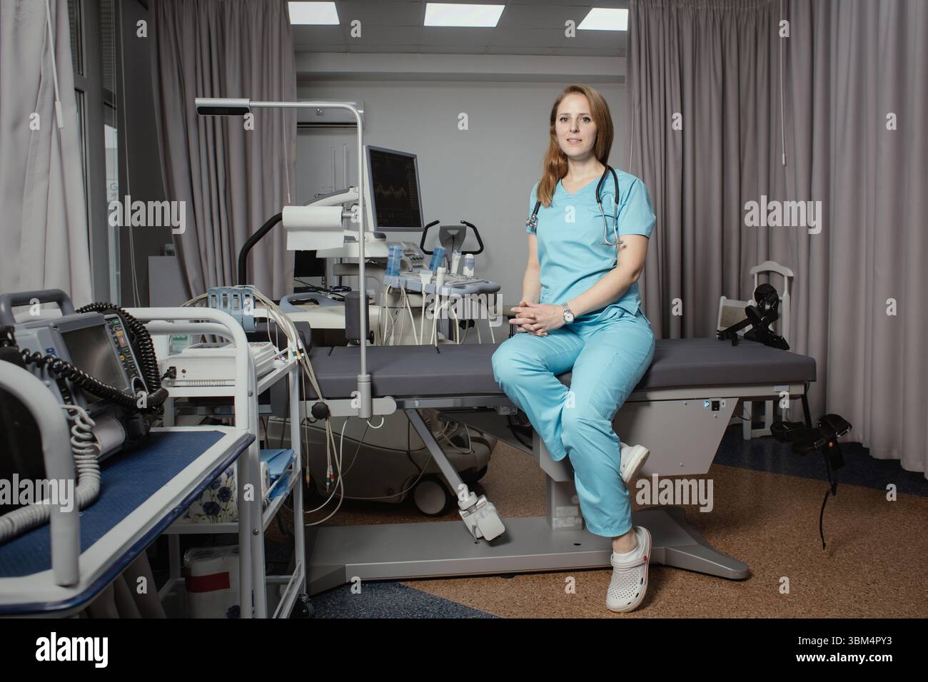 Ritratto di donna medico o ecografo in scrub in piedi nella moderna sala diagnostica a ultrasuoni con attrezzature mediche e stetoscopio. Foto Stock