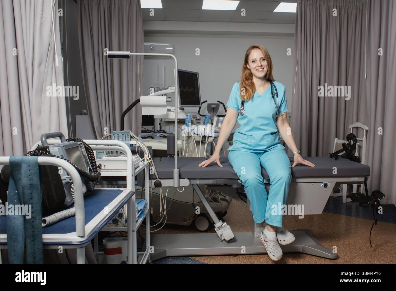Ritratto di donna medico o ecografo in scrub in piedi nella moderna sala diagnostica a ultrasuoni con attrezzature mediche e stetoscopio. Foto Stock