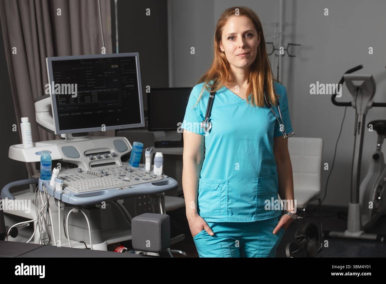 Ritratto di donna medico o ecografo in scrub in piedi nella moderna sala diagnostica a ultrasuoni con attrezzature mediche e stetoscopio Foto Stock