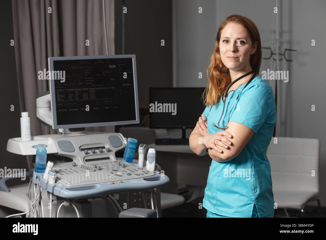 Ritratto di donna medico o ecografo in scrub in piedi nella moderna sala diagnostica a ultrasuoni con attrezzature mediche e stetoscopio Foto Stock