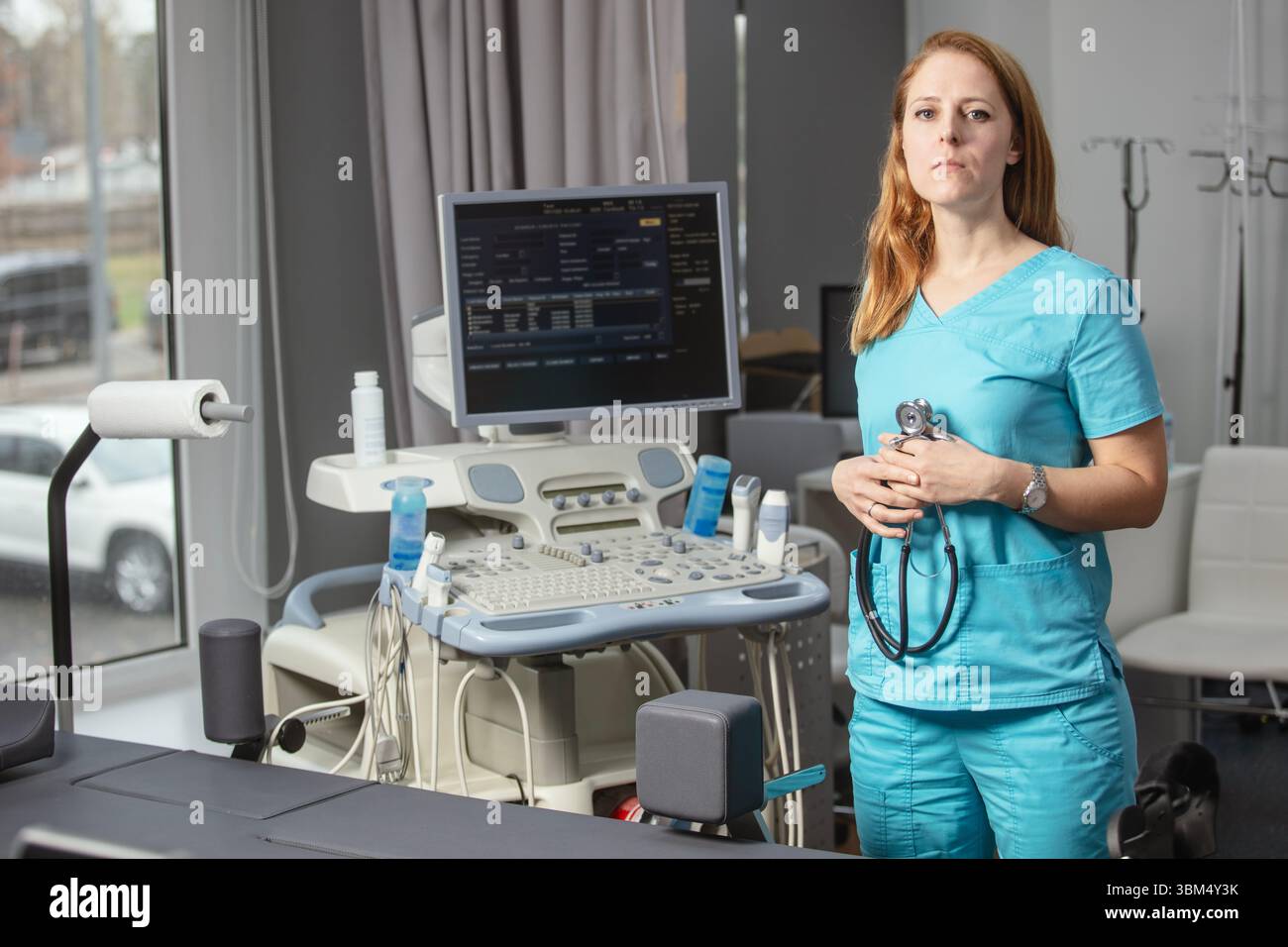 Ritratto di donna medico o ecografo in scrub in piedi nella moderna sala diagnostica a ultrasuoni con attrezzature mediche e stetoscopio Foto Stock
