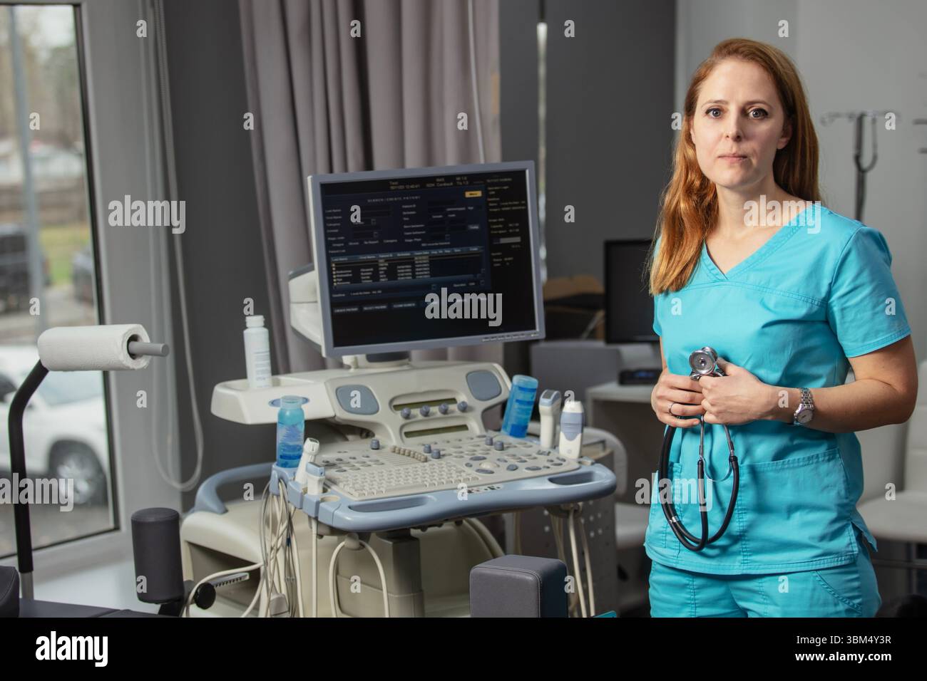 Ritratto di donna medico o ecografo in scrub in piedi nella moderna sala diagnostica a ultrasuoni con attrezzature mediche e stetoscopio Foto Stock