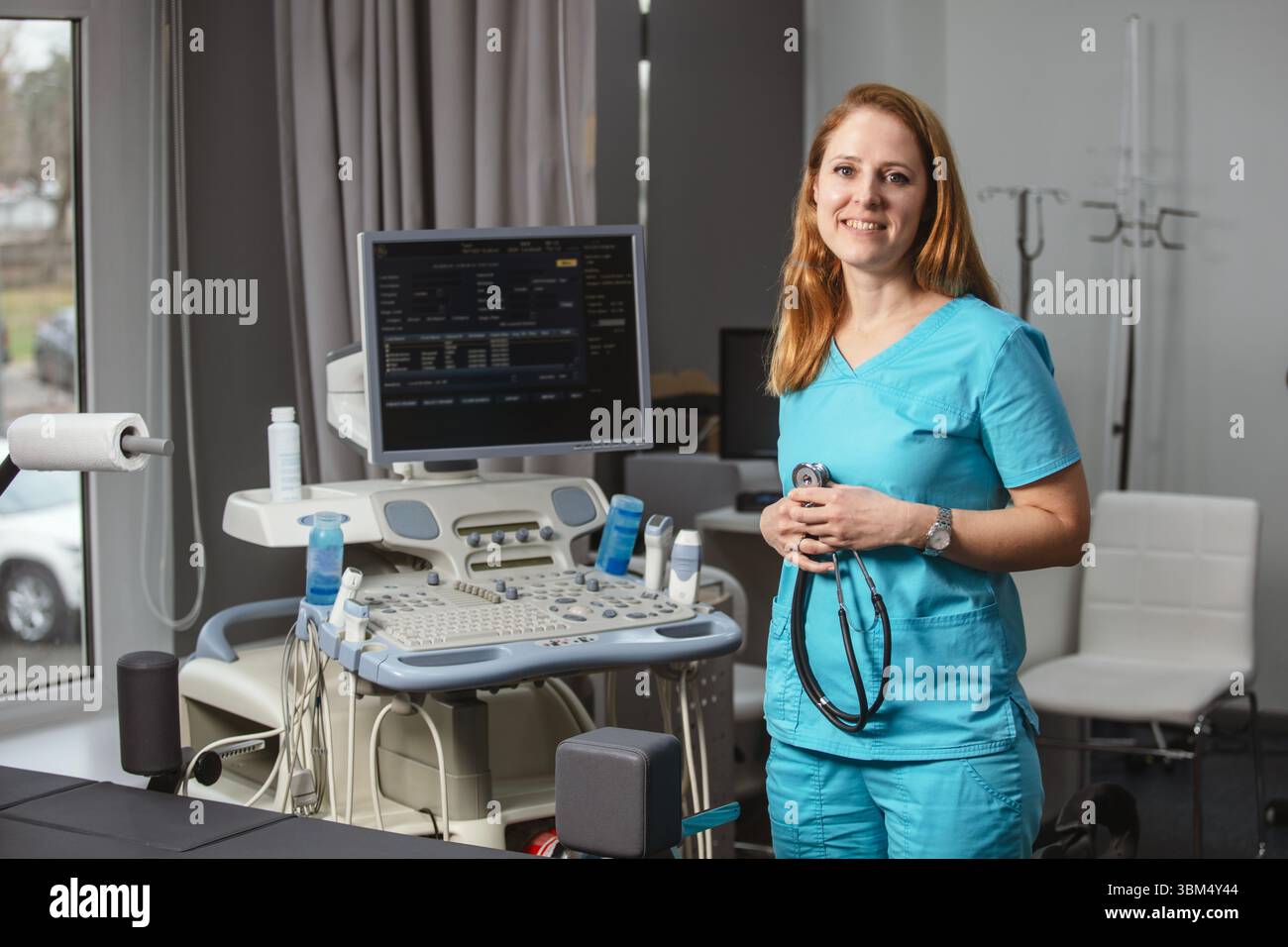 Ritratto di donna medico o ecografo in scrub in piedi nella moderna sala diagnostica a ultrasuoni con attrezzature mediche e stetoscopio Foto Stock