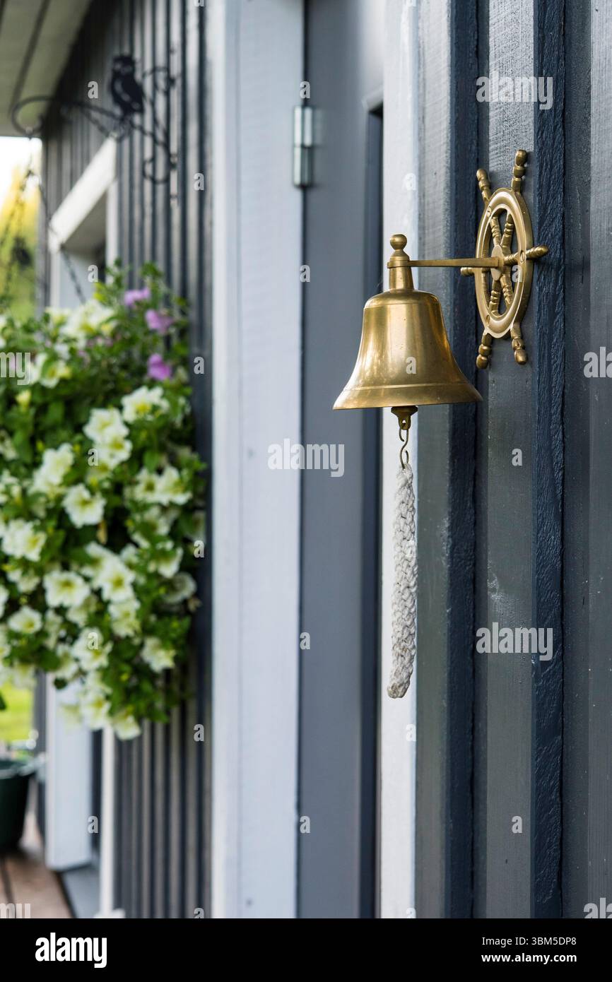 Campana in ottone montata a parete in stile nautico su una base a forma di ruota della nave, utilizzata come campanello, con vasi di fiori appesi sullo sfondo. Foto Stock