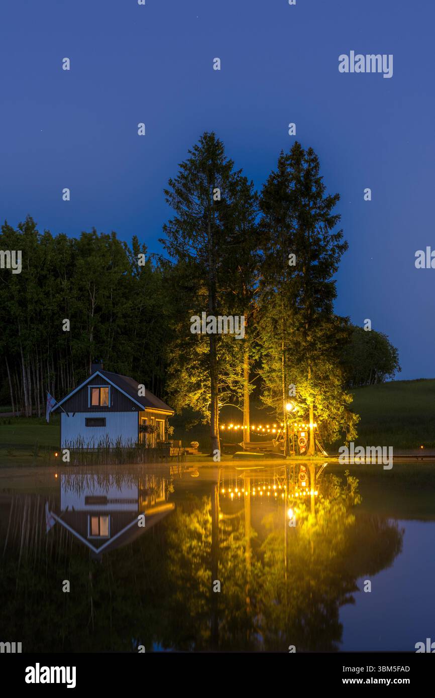 Un'accogliente cabina sul lago nell'Estonia rurale illuminata da calde luci all'aperto, riflessa nell'acqua calma in una tranquilla notte d'estate. Foto Stock