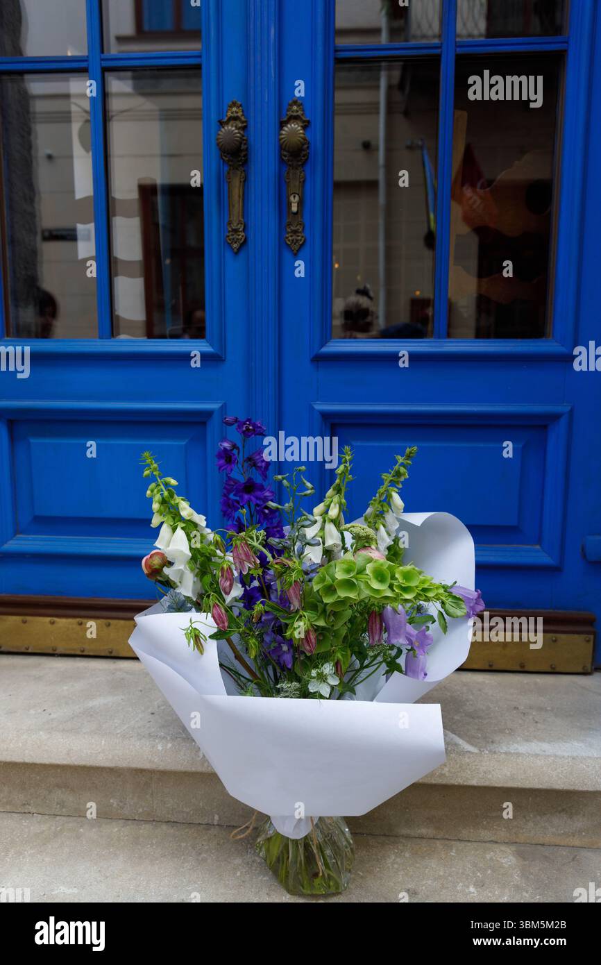 Questa immagine mostra un pittoresco bouquet di fiori variegati elegantemente posto ai piedi di un audace ingresso blu a doppia porta, che mette in risalto la bellezza Foto Stock
